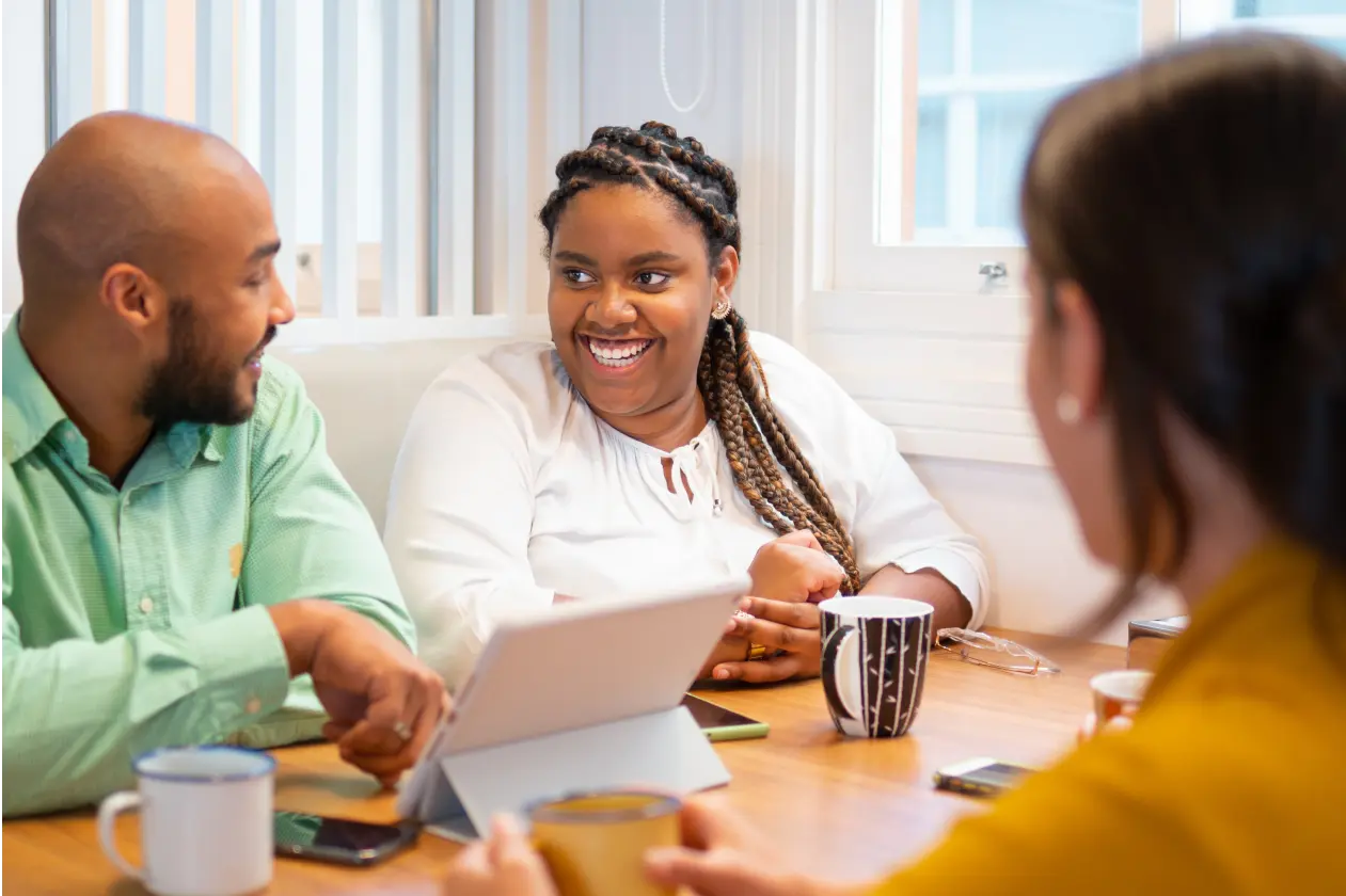 Um homem de pele negra, cabeça raspada, barba curta e usando camisa verde social, uma mulher de pele negra, cabelos escuros com tranças e usando camisa branca de manga longa e uma mulher de pele clara e cabelo escuro usando camisa amarela, de costas. Todos sentados à mesa de um escritório, sorrindo e conversando.