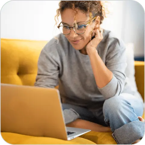 Mulher parda, cabelos cacheados com mechas loiras, preso, usando óculos azul, camisa cinza de manga longa e calça jeans clara, sentada num sofá amarelo, utilizando um notebook sorrindo.