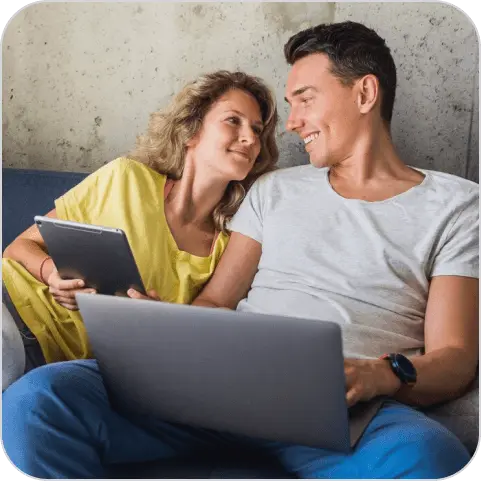 Mulher com cabelo loiro e solto, usando camisa amarela, segurando um tablet, sentada num sofá azul sorrindo e olhando para um homem ao lado dela de pele clara, cabelo preto curto, usando camisa cinza, segurando um notebook, também sorrindo.